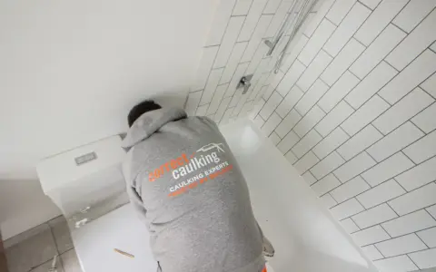 A person in a "Correct Caulking" hoodie works on a bathtub, demonstrating how to caulk a bathtub with white subway tiles in the background.