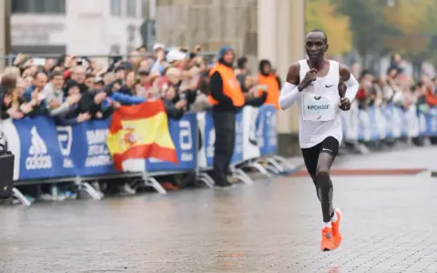 Eliud Kipchoge corre in una maratona, mostrando quanto è lunga una maratona. Pubblico festante e bandiera spagnola sullo sfondo.