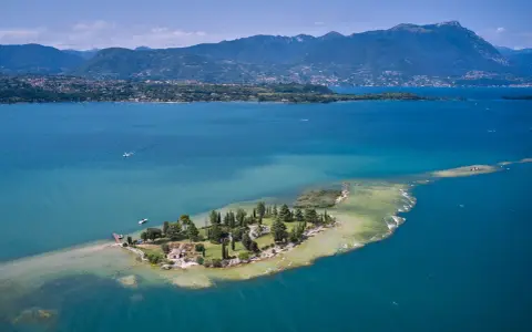 L'isola dei conigli sul Lago di Garda, un gioiello verde con case e alberi, circondata da acque turchesi e montagne sullo sfondo.