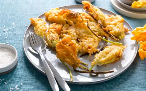 Piatto di fiori di zucca in pastella dorati e croccanti, serviti con sale grosso e posate.