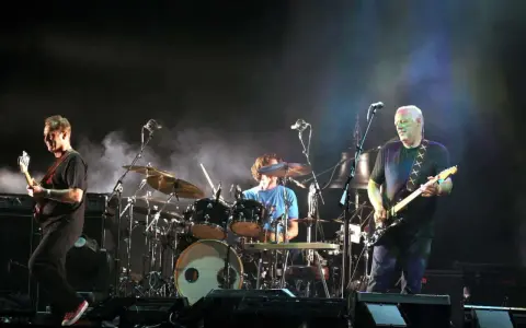 Les membres de Pink Floyd sur scène, David Gilmour à la guitare, Nick Mason à la batterie, et un autre musicien.