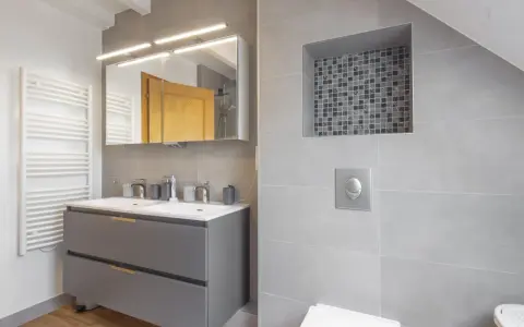 Salle de bain moderne avec double vasque, radiateur blanc et un choix carrelage gris subtil, agrémenté d'une niche décorative en mosaïque.
