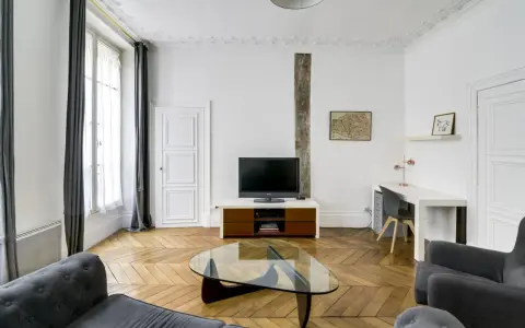 Salon moderne avec parquet en chevrons, canapés gris, bureau et TV. Les murs et le plafond sont peints à l'huile, offrant une finition impeccable.