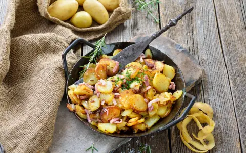 Bratkartoffeln, die vorher gekocht wurden, in einer Pfanne mit Speck und Kräutern garniert. Daneben liegen rohe Kartoffeln in einem Jutesack.