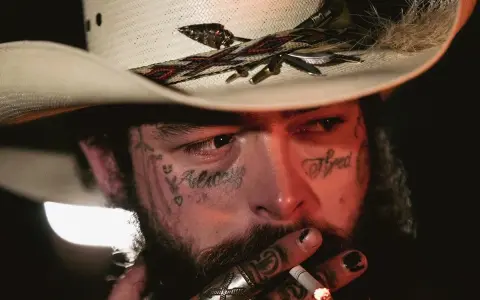 Amerykańskie gwiazdy muzyki country, jak Post Malone, w kowbojskim kapeluszu, z tatuażami na twarzy, palący papierosa.