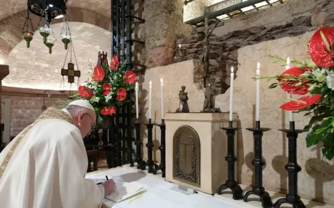 Papst Franziskus schreibt in einem Buch, während Kerzen und rote Anthurien brennen. Dies erinnert an die Bedeutung einer Enzyklika, eines wichtigen päpstlichen Schreibens.