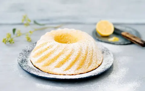 En saftig sockerkaka citron, pudrad med florsocker, serveras på ett fat. En halv citron och en kniv syns i bakgrunden.