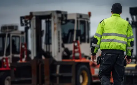 Arbetare i varseljacka inspekterar en truckladdning med flera gaffeltruckar i bakgrunden.