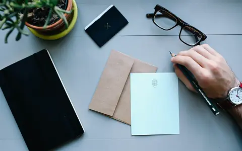 Ręka pisze ołókiem na jasnoniebieskiej papeterii, przygotowując list do Anglii. Obok zeszyt, okulary i koperta.