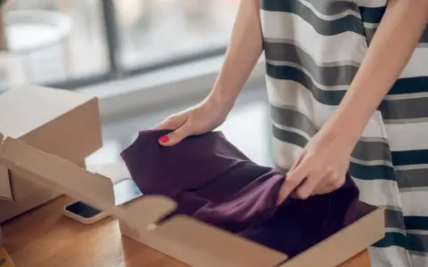 Kobieta pakuje ciemnofioletową sukienkę do kartonu, przygotowując ją do **vinted zwroty**.