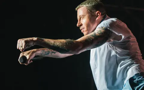 Macklemore na koncercie w Polsce, z mikrofonem w dłoni, w białej koszuli i z tatuażami na ramionach.
