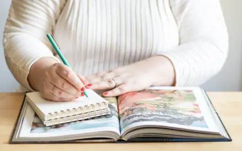 Frau schreibt mit grünem Stift in Notizbuch, während sie ein aufgeschlagenes Fotoalbum betrachtet. Sie erstellt ihr persönliches Jahrbuch.