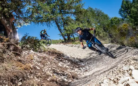 Due ciclisti affrontano un sentiero sterrato in discesa, immersi nella natura. La **tonezza del Cimone** si percepisce nell'aria fresca e nel paesaggio montano.