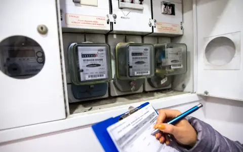 Kontrola liczników energii elektrycznej. Ręka z długopisem zapisuje dane na kartce, obok widoczne liczniki i bezpieczniki.