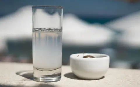 Vaso de pastis bebida, con una aceituna en un cuenco blanco. El fondo está desenfocado, sugiriendo un día soleado.