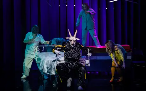 Na scenie, w surrealistycznej atmosferze, panna obsada w stroju z głową kozła siedzi na wózku. Wokół niej, w jasnych fartuchach, poruszają się postacie przypominające personel medyczny.