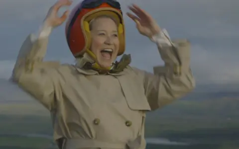 Glad Trine Dyrholm i pilotmössa och trenchcoat vinkar glatt. En scen från filmer med Trine Dyrholm.