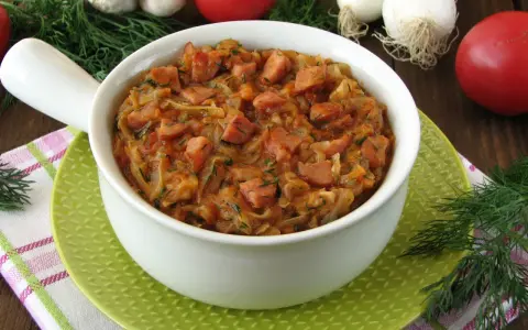 Pyszny bigos z młodej kapusty z kiełbasą, podany w białej misce na zielonym talerzyku. W tle pomidory, czosnek i koperek.