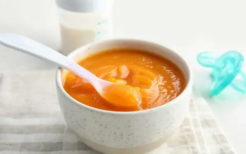 Un tazón con puré de calabaza bebé, una cuchara rosa y un biberón.