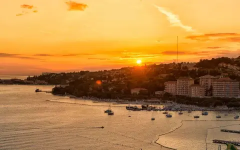 Tramonto dorato sul mare in Slovenia, con un porto pieno di barche e una costa verdeggiante. Immagina i bambini che giocano felici sulla spiaggia.
