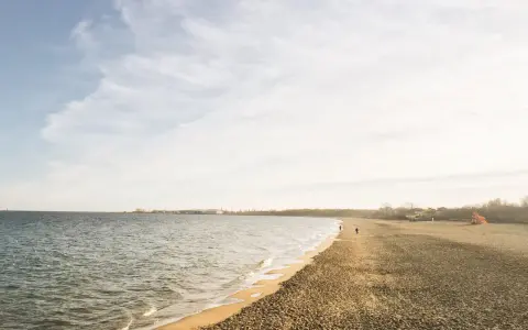 Spokojny brzeg jeziora Białe Kaszuby, fale delikatnie muskają piaszczysty brzeg, a na horyzoncie widać zarys zabudowań.