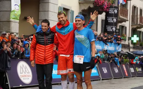 Trzech biegaczy na podium po ultra-trail du mont-blanc. Jeden w pomarańczowej kurtce, drugi w czerwonej, trzeci w niebieskiej koszulce.