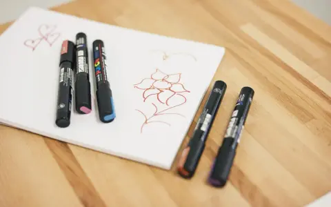 Några Posca-pennor ligger på ett vitt papper med en enkel blomma och hjärtan ritade i rött.