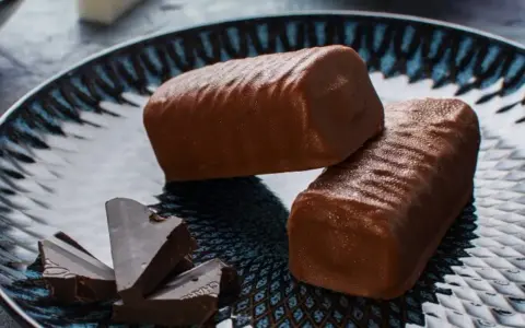 Dwa czekoladowe batony proteinowe na talerzu, obok kawałki gorzkiej czekolady. Idealne do domowego batony proteinowe przepis.