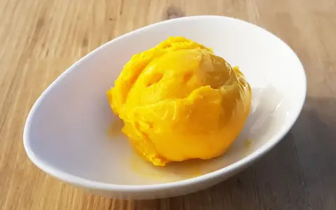 Ein leuchtend gelber Löffel Mango Sorbet in einer weißen Schale auf Holzhintergrund.