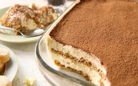 Pyszne tiramisu bez jajek w szklanej formie, posypane kakao. Obok porcja deseru na talerzyku.