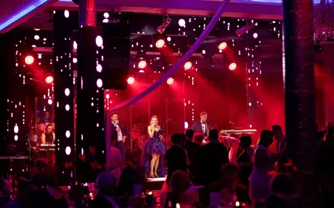 Sylwester w Hotelu Gołębiewski: zespół na scenie, wokalistka w fioletowej sukni, muzycy i publiczność bawią się przy czerwonych światłach.