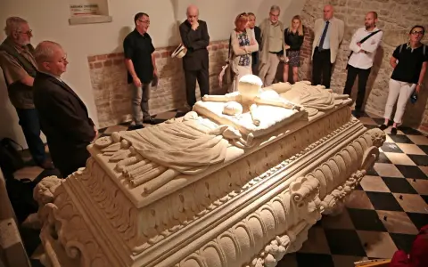 Grupa zwiedzających ogląda kamienny sarkofag w wawel krypty. Kamienne rzeźbienia zdobią jego bok.
