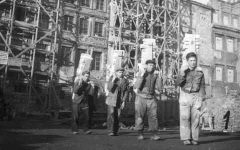 Mężczyźni niosą cegły na tle odbudowywanej kamienicy. To obraz Warszawy po wojnie, gdzie trwa mozolna praca nad przywróceniem miasta do życia.