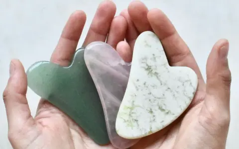 Drei herzförmige Gua Sha Steine in Grün, Rosa und Weiß liegen in einer Hand. Ideal für Gua Sha gegen Falten.