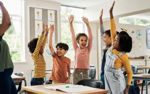Fröhliche Kinder mit erhobenen Armen in einem Klassenzimmer. Sie nehmen begeistert an Kennenlernspielen für die Grundschule teil.