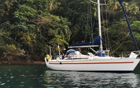 Un voilier blanc, un voilier Jurançon, est ancré dans une baie tropicale luxuriante, entouré d'une végétation dense et de palmiers.