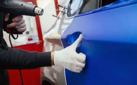 Ein Handwerker erwärmt mit einem Heißluftföhn eine blaue Folie, die auf ein Auto aufgebracht wird. Die auto folieren kosten sind hierbei ein wichtiger Faktor.