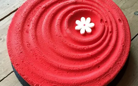 Un tiramisu fraise rhubarbe d'un rouge éclatant, décoré d'une petite fleur blanche en son centre, sur un fond de bois.
