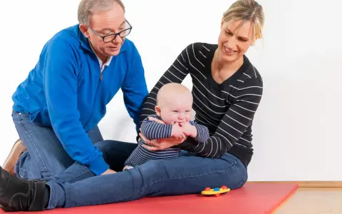 Ein Mann und eine Frau üben mit einem Baby auf einer roten Matte. Das Baby greift nach seinen Fingern. kg zns kinder.