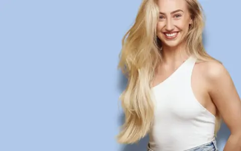 En leende kvinna med långt, blont hår som ser ut att bleka håret naturligt. Hon bär en vit topp och jeans mot en ljusblå bakgrund.