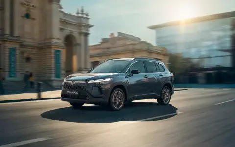 Ciemnoszary Toyota Corolla Cross Hybrid sunie ulicą miasta, odbijając światło słoneczne od nowoczesnych budynków.