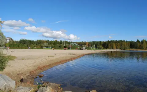 Solig strand vid Nydala camping Umeå med klart vatten och grönskande skog i bakgrunden.