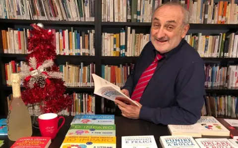 Daniele Novara, autore di libri per genitori, legge un suo libro tra scaffali pieni di volumi, con un albero di Natale e una bottiglia di spumante.