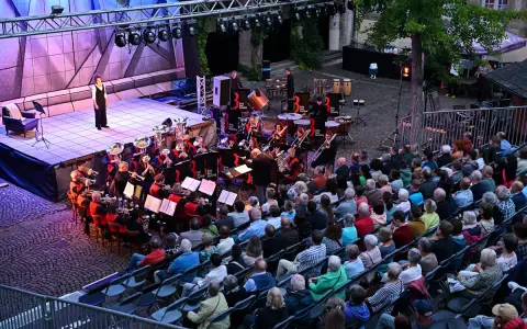 Eine Brass Band spielt auf einer Bühne, während ein Publikum auf Stühlen sitzt und zuhört.