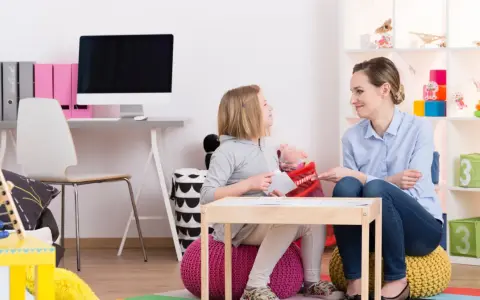 Terapeutka pracuje z dziewczynką, wspierając ją w terapii afazji u dzieci. W tle widać pomoce dydaktyczne i zabawki.
