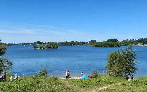 Sonniger Tag am Lippesee Paderborn. Menschen entspannen am Ufer, Segelboote gleiten über das Wasser, eine kleine Insel ziert die Mitte.