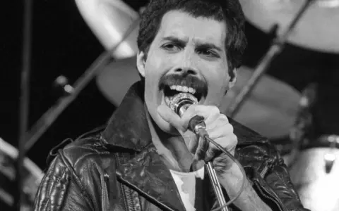 Freddie Mercury przed śmiercią, w skórzanej kurtce, śpiewa do mikrofonu. W tle widać perkusję.