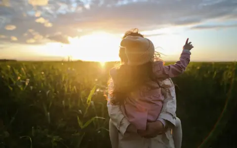Bambina aggrappata alla schiena di un adulto, con il sole al tramonto che illumina un campo. Un'immagine di puro attaccamento affettivo.
