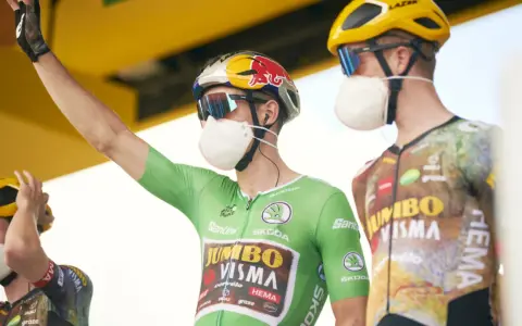 Cyclist in a green jersey, the symbol of the points classification leader at the Tour de France, raises his arm. He wears a mask and helmet.