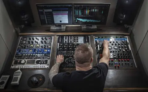 Un homme aux bras tatoués ajuste les réglages d'une console de mixage audio, entouré d'écrans affichant des formes d'ondes et des analyseurs.